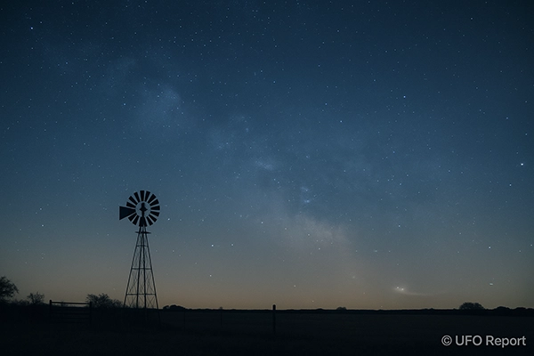 A quiet Texas field at night under a starlit sky, with a faint circular glow or light trail near the horizon.