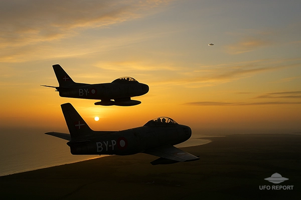 Two Danish fighter jets banking over a coastline at sunrise. In the distance, a small reflective disc-shaped glint appears high above the horizon, barely noticeable but clearly out of place.